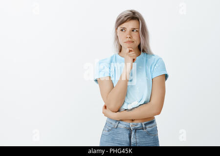 Girl feeling not sure about received suggestion. Doubtful intense charming woman with fair hair, frowning and pursing lips while holding hand on chin and looking aside, thinking, making choice Stock Photo