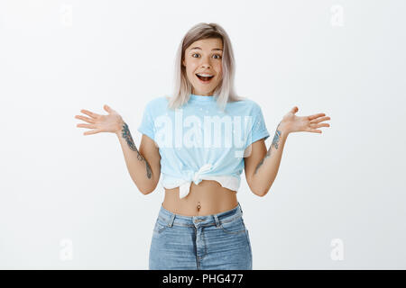 What great surprise. Portrait of happy emotive young woman with fair hair tattoos and pierced belly, smiling broadly and spreading raised palms, being astonished with unexpected meeting with friend Stock Photo