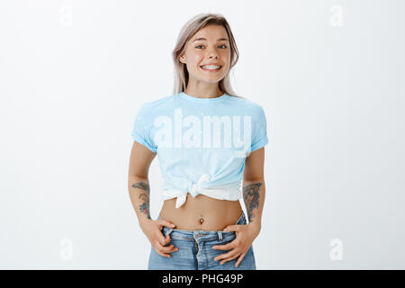 Portrait of joyful good-looking girlfriend with fair hair and tattoos, holding hands in pockets and smiling joyfully, showing cute and shiny braces, picking new clothes in shop with friend Stock Photo