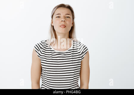 Indoor shot of playful good-looking woman in striped t-shirt, tilting head and showing tongue, being childish and in great mood, playing with friend and making faces while being bored during classes Stock Photo