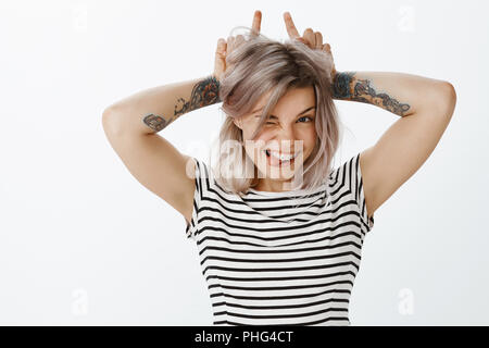 Cute girl little devilish inside. Portrait of cute playful woman with fair hair, braces and tattoos, showing horns with index fingers on head, winking and smiling while sticking out tongue Stock Photo