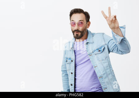 Peace folks. Portrait of middle-aged handsome caucasian man with dark hair and beard, raising hand with peace gesture, smiling and folding lips, being high and having fun on disco party Stock Photo