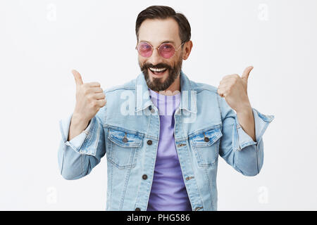 Yeah I like it. Pleased and satisfied good-looking male in sunglasses and denim jacket, raising thumbs up and smiling broadly, giving positive feedback about party club, standing joyful over gray wall Stock Photo