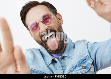 Wats up. Carefree excited and joyful man acting like teenager in sunglasses and denim jacket, pulling hands towards camera, taking selfie, smiling joyfully, sticking out tongue and squinting from joy Stock Photo