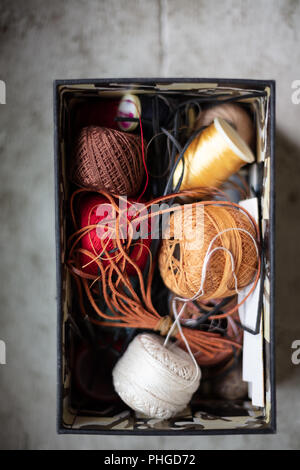 Close up view of assorted twines, string and thread rolls in an open container viewed from overhead on a rustic wood shelf below a window under sunshi Stock Photo