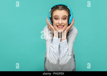 Happy girl in striped blue shirt and pigtail hairstyle, standing listening music and holding her headphones and looking at camera with toothy smile, I Stock Photo