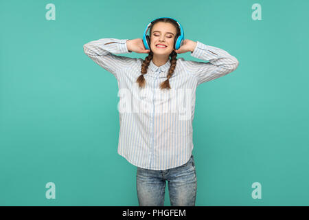 Happy satisfied girl in striped blue shirt and pigtail hairstyle, standing listening favorite music with headphones, closed eyes and toothy smiling, I Stock Photo