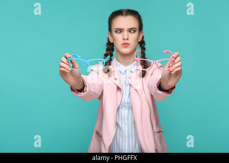 doubtful funny girl in casual or hipster style, pigtail hairstyle, standing, holding blue and pink glasses and looking at camera with crossed eye, Ind Stock Photo