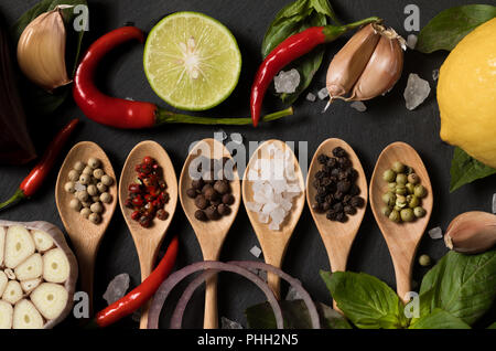 Different kind of pepper and salt with garlic and basil leaf in wooden spoons on a black slate background Stock Photo