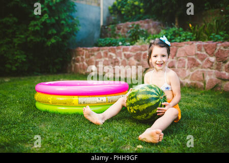 The topic proper healthy eating children. Little baby girl 4 years old caucasian sitting on green grass hugging big green round berries watermelon nea Stock Photo