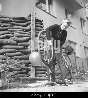 1940s woman on a bicycle. Swedish actress Viveca Lindfors is repairing her bicycle. Notice the sand bags placed in front of the apartment building and the entrance to the basement shelter.  Sweden August 1940. Photo Kristoffersson 159-32 Stock Photo