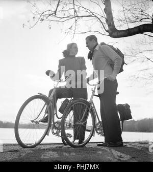 1940s couple on bicycles. A young couple is out on a tour on their bicycles on a sunny spring day. They have practical bags attached to the racks of the bicycles where they could transport the picnic food.  Sweden 1947. Photo Kristoffersson AB11-10 Stock Photo