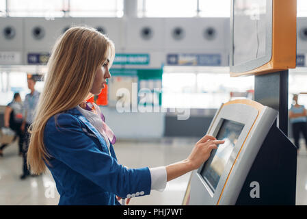 Slender stewardess uses the terminal at the airport hall. Air hostess with baggage in departure area, flight attendant with hand luggage, aviatranspor Stock Photo