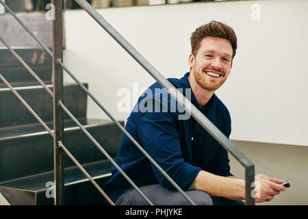 Smiling young businessman sitting on stairs in an office reading text messages on a cellphone Stock Photo