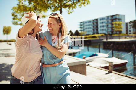 Two female friends laughing while walking arm in arm together along a city promenade in summer Stock Photo