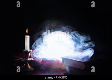 Crystal ball for wizard and fortune teller smokes and lights on table and candle in candlestick and old books, black background. Halloween concept Stock Photo