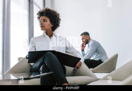 Young businesswoman sitting on office lounge with a file and male colleague working in background. Female executive looking away while working in offi Stock Photo