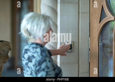 Slightly blurry foreground woman pressing black button on doorway wall for doorbell in front of glass and wooden door Stock Photo