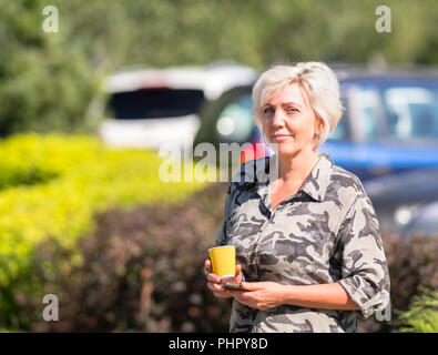 Confident woman with white hair holding small orange cup and smart phone while blurry cars sit in background Stock Photo