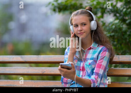 Young woman relaxing on an outdoor bench listening to music on stereo headphones looking to the side with a thoughtful expression Stock Photo