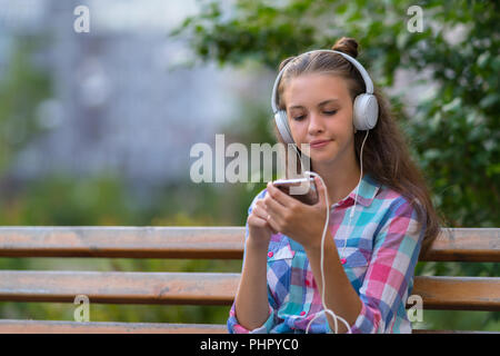 Young woman trying to decide on a new soundtrack looking at her mobile phone with a thoughtful expression as she listens to music outdoors Stock Photo