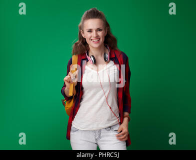 Portrait of smiling young student woman in a red shirt with backpack and headphones against green background Stock Photo