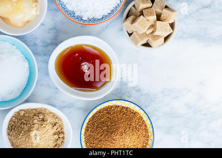 Different Kinds of Sugar and Sweeteners in the Bowls, such as coconut, pure cane, icing, maple syrup, dark brown soft sugar, honey, demerara cubes Stock Photo