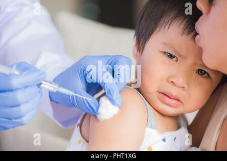 Doctor giving an injection vaccine to a girl. Little girl crying with her mother on background. Stock Photo