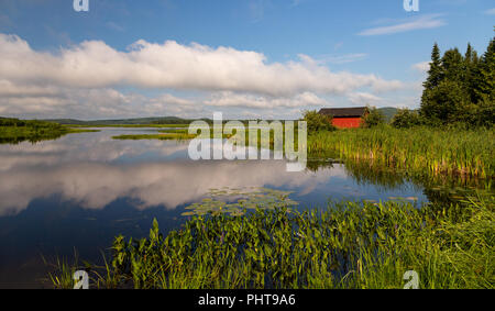 Tranquil landscape shot of a small red fishing shed on Eel River, Benton, New Brunswick, Canada. Fluffy white clouds reflected in the water. Stock Photo