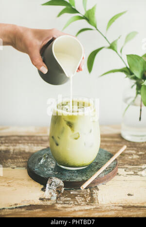 Iced matcha latte drink in glass with coconut milk pouring from pitcher by woman's hand, white wall and plant branches at background. Summer refreshin Stock Photo