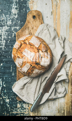 Flat-lay of freshly baked sourdough wheat bread loaf on board over linen napkin and rustic wooden tray background, top view, vertical composition Stock Photo
