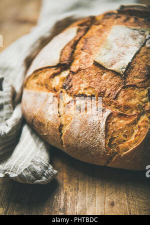 Freshly baked sourdough wheat bread loaf over linen napkin and rustic wooden table background, selective focus, vertical composition Stock Photo