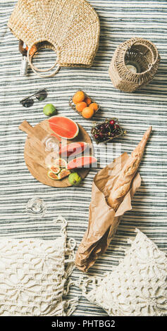 Summer picnic setting. Rose wine in glass and bottle, fresh fruit on board, baguette on blanket and woman's straw bag, top view. Outdoor gathering or