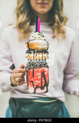 Young woman in light pink clothing holding summer cold strawberry donut freakshake drink with whipped cream in mason jar in hand, selective focus, ver Stock Photo