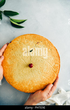 A woman holding a cherry pie topped with a single cherry photographed from top view. Green leaves and white linen on grey background accompany. Stock Photo