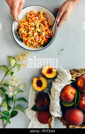 A woman stirring peach salsa in a white bowl photographed from top view. Fresh peaches in a basket and tiny flowers with green leaves accompany. Stock Photo
