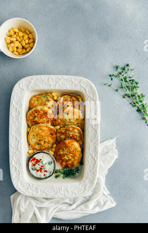 Corn fritters served with yogurt sauce and fresh thyme on the side in a white pan photographed on a grey background from top view. Fresh thyme sprigs Stock Photo