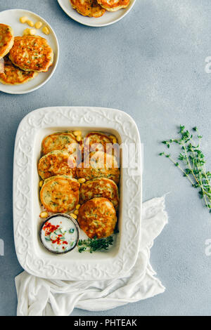 Corn fritters served in a white pan with a small bowl of yogurt sauce on the side and on two white plates photographed on a grey background from top v Stock Photo