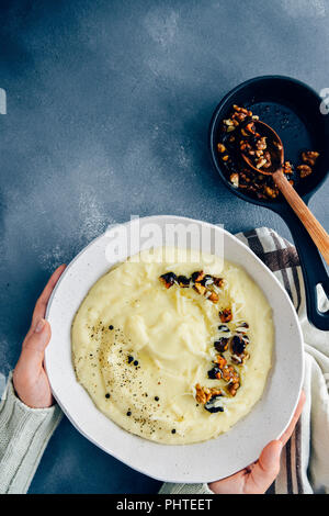 Hands holding cheesy mashed potatoes with roasted walnuts and black pepper in a white ceramic bowl photographed on a dark background from top view. Stock Photo