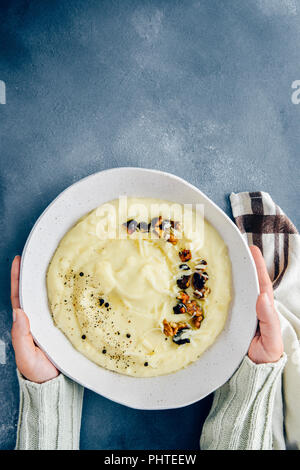 Hands holding cheesy mashed potatoes with roasted walnuts and black pepper in a white ceramic bowl photographed on a dark background from top view. Stock Photo