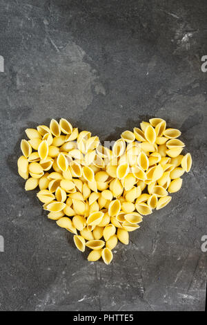 Raw macaroni - pasta Conchiglie. In the middle of the empty space in the shape of a heart on a dark concrete background Stock Photo