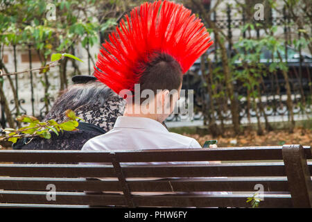 A punk with a very large scarlet mohican and a safety pin earring sits on a park bench in London Stock Photo