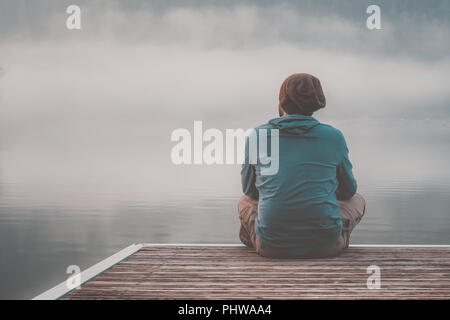 Young man with hood sitting on the jetty of a lake on a foggy day.