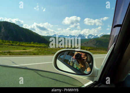 Reflection in the mirror of a riding white car of photographer who makes photo of a large altai mountains with snow on top by a sunny bright day with  Stock Photo