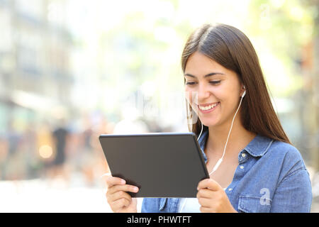 Happy girl wearing earphones watching streaming videos online in a tablet in the street Stock Photo