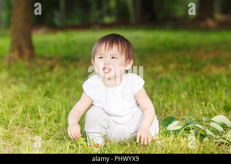 Portrait of cute adorable little Asian girl child baby one year old in white pants shirt, standing on field meadow grass on sunset playing Stock Photo