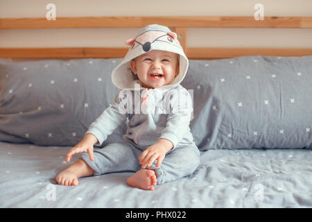 Portrait of cute adorable Caucasian blonde smiling baby girl with blue eyes in grey pajama with fox cat animal hood sitting on bed in bedroom. Natural Stock Photo