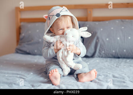 Portrait of cute adorable Caucasian blonde smiling baby girl with blue eyes in grey pajama with fox cat animal hood sitting on bed in bedroom and hold Stock Photo