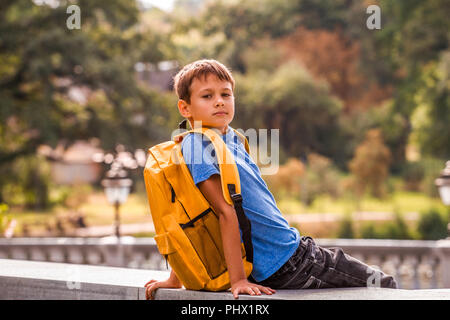 Cute smart boy with backpack sitting outdoors after school. Stock Photo