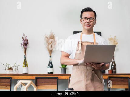 Asian male Barista cafe owner using laptop in cafe business inside coffee shop,looking and smile at camera.food and drink business start up Stock Photo